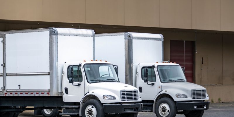 Two Industrial standard professional white day cab middle duty rig semi truck tractors with box trailers standing on the warehouse dock gate loading cargo for the next commercial delivery